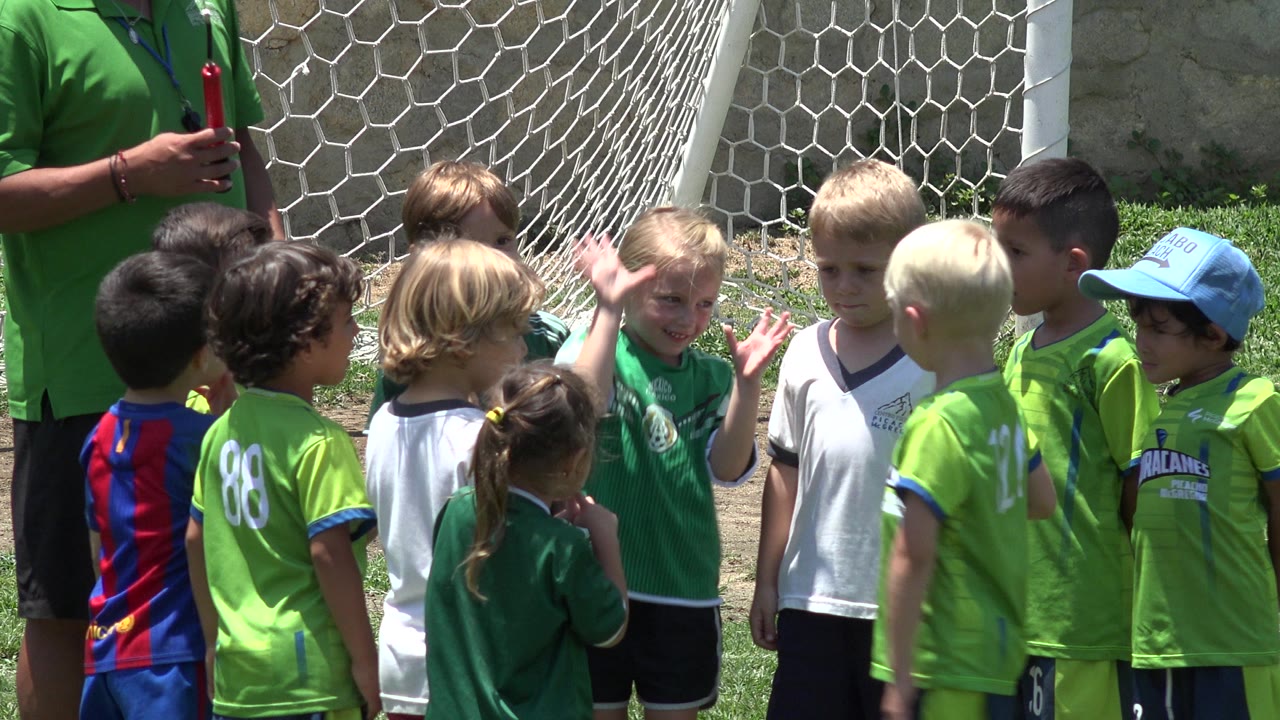A child in a green jersey raises a hand while teammates gather around the goal net in Cabo San Lucas, Mexico, with the coach still partly cropped at left.