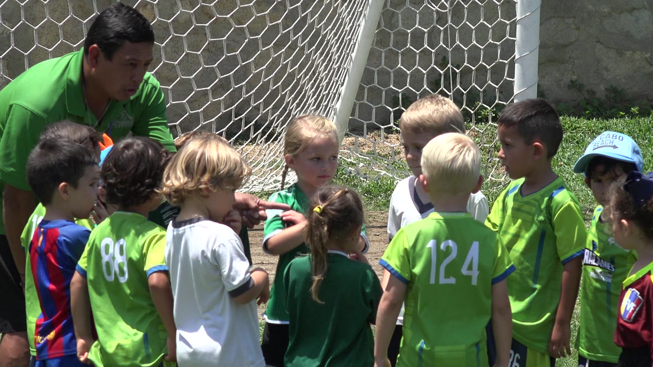 Children in green soccer jerseys stand near a white goal net with an adult leaning in.