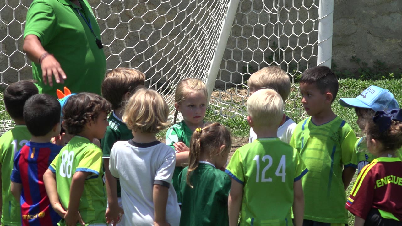 Children in green football shirts gather beside a goal net with a coach partly visible.