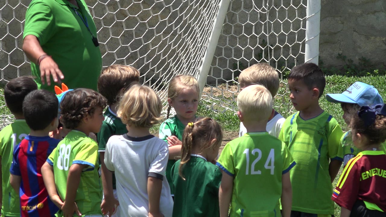 A group of children in bright football shirts stand in front of the goal net in Cabo San Lucas, Mexico as the cropped coach gestures overhead.