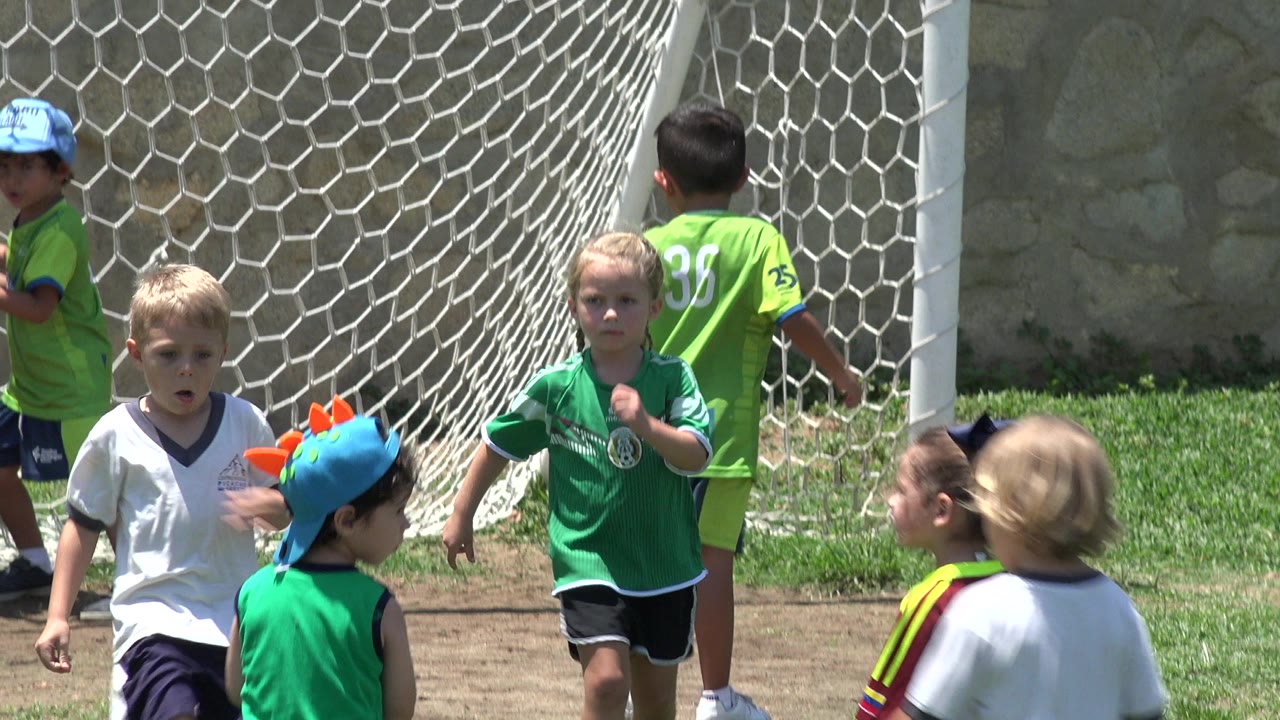 Several young children in soccer jerseys stand near a white goal net on a sunny field in Cabo San Lucas, Mexico, with grass, dirt, and a stone wall behind them.