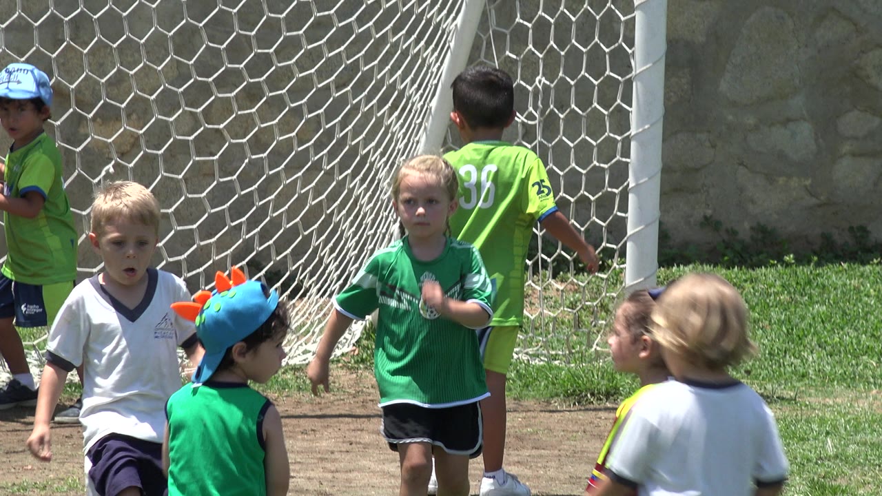 Young children in green and white soccer tops move in front of a white goal net in Cabo San Lucas, Mexico, while another child stands with their back to camera.