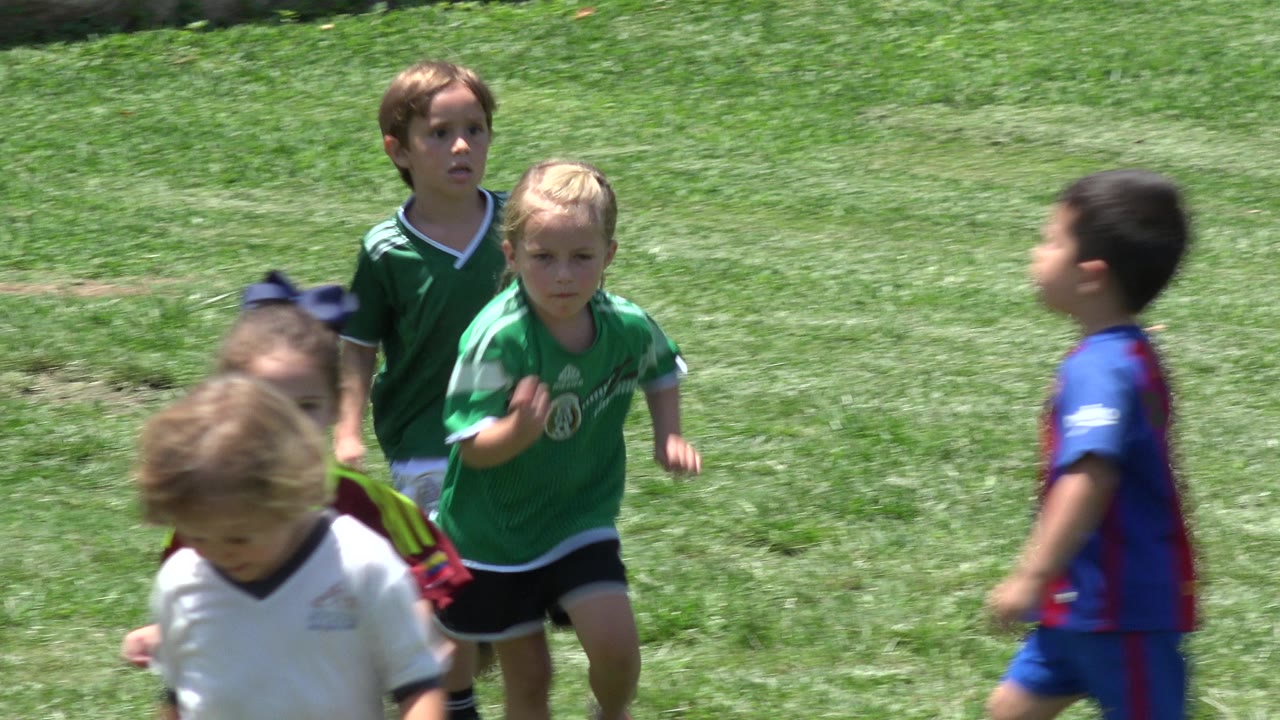 Young kids continue running over the manicured grass in Cabo San Lucas, Mexico, with green jerseys in the center and a blue-red jersey crossing at right.