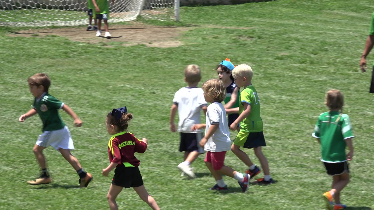 Young children in soccer jerseys run across a bright grass field in Cabo San Lucas, Mexico, with a small goal and fence behind them.