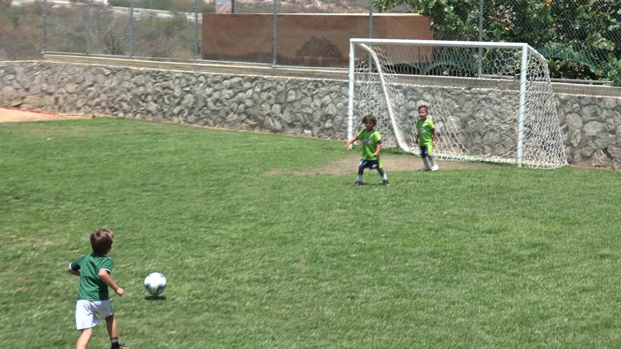 At 0:00 in Cabo San Lucas, Mexico, a child in a green shirt runs toward a soccer ball while two children stand near a white goal on a grass field.