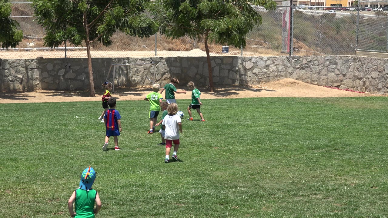 Children play soccer on a sunlit grass field in Cabo San Lucas, Mexico, with a child in a green shirt and helmet-like cap in the foreground and several players near the wall.