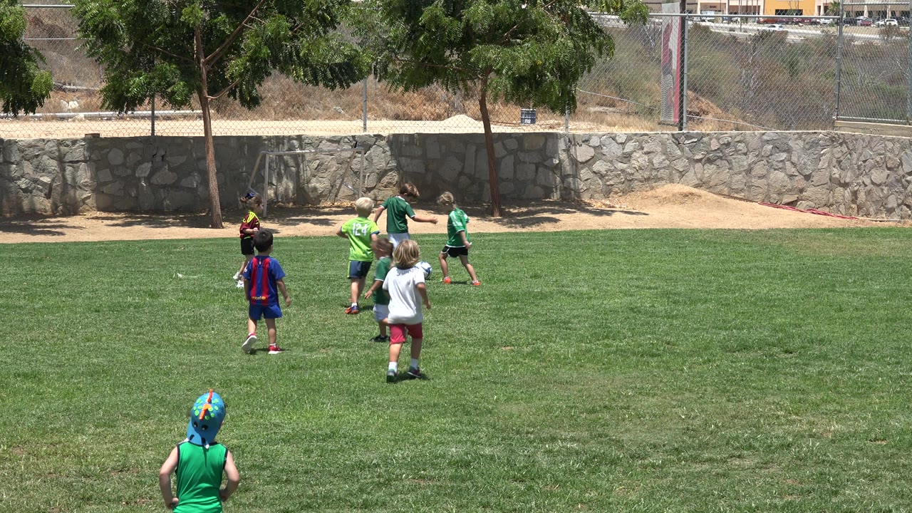 Several children in green jerseys and casual sports clothes move across the Cabo San Lucas grass field, with the stone wall, trees, and fence holding steady behind them.