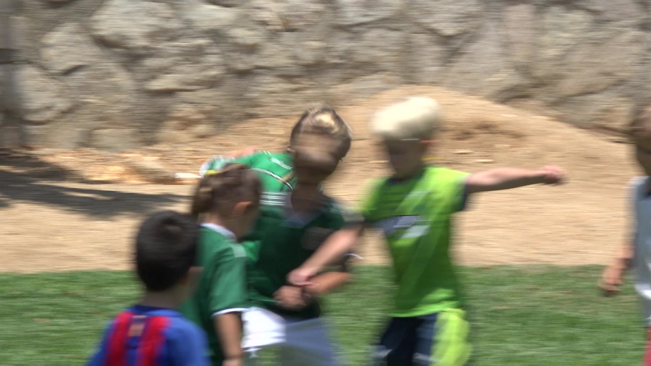 A tight group of children runs through the soccer action in Cabo San Lucas, Mexico, with a lime shirted child blurred at the right and a striped shirt visible in the foreground.
