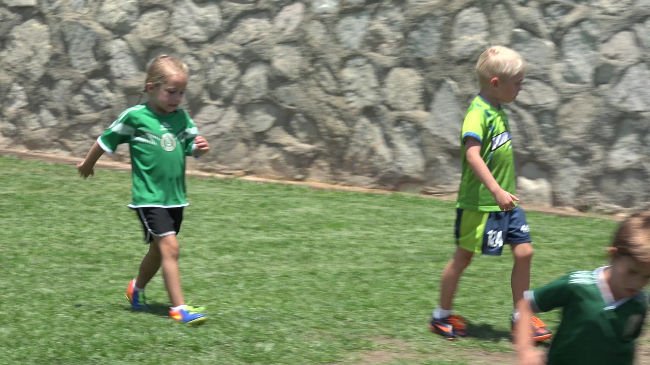 A child in a green soccer jersey moves across a sunny grass field in Cabo San Lucas, Mexico, with a stone wall behind her.