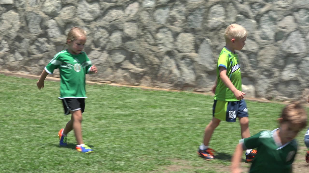 A blond child in a bright green soccer kit stands on the grass in Cabo San Lucas, Mexico, with the stone boundary wall filling the background.