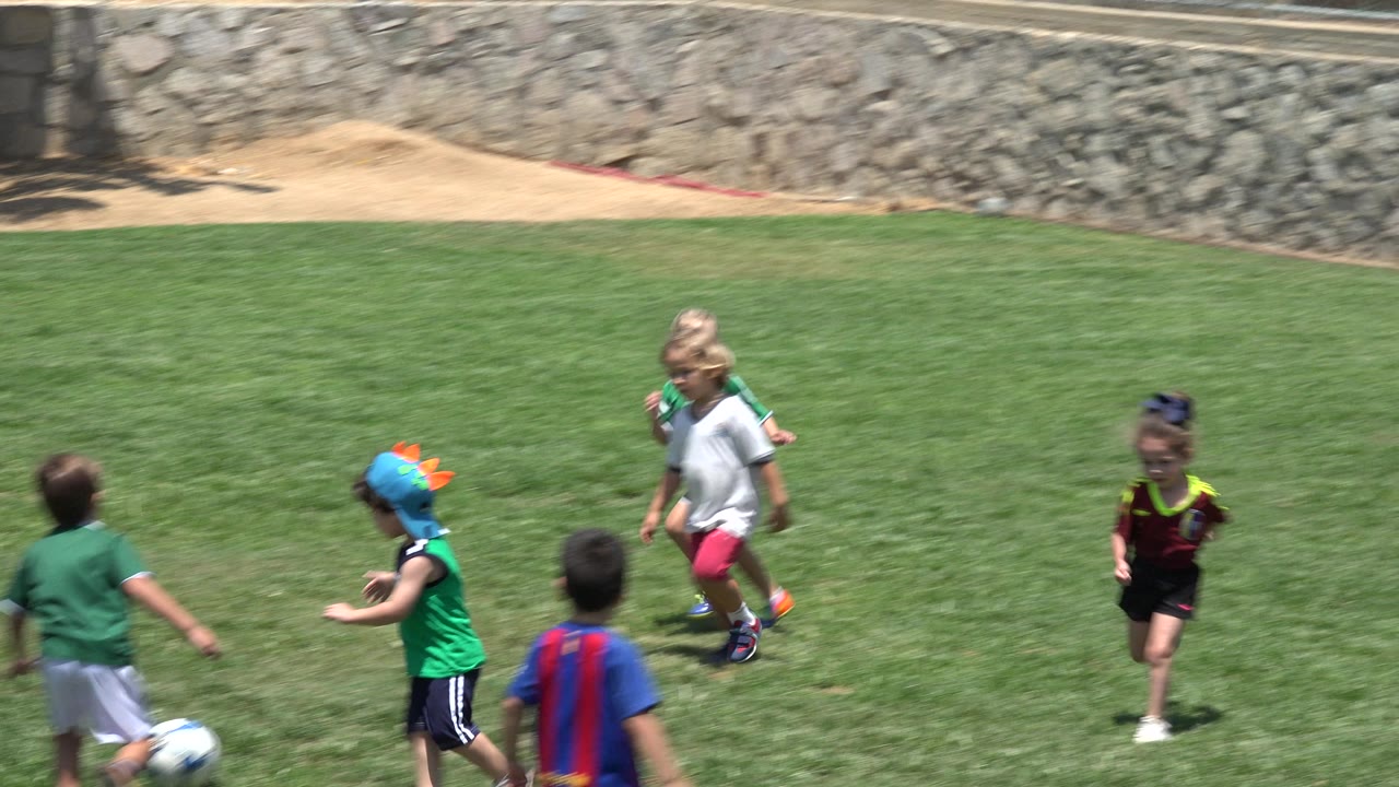 Children in green soccer uniforms run across a grass field near a ball.