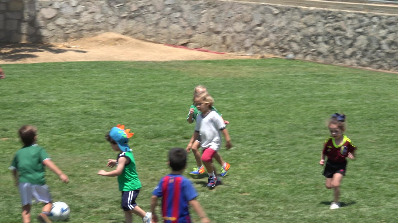 A group of children in Cabo San Lucas runs across the grass field, including green uniforms, a white shirt, and a blue and red jersey in the foreground.