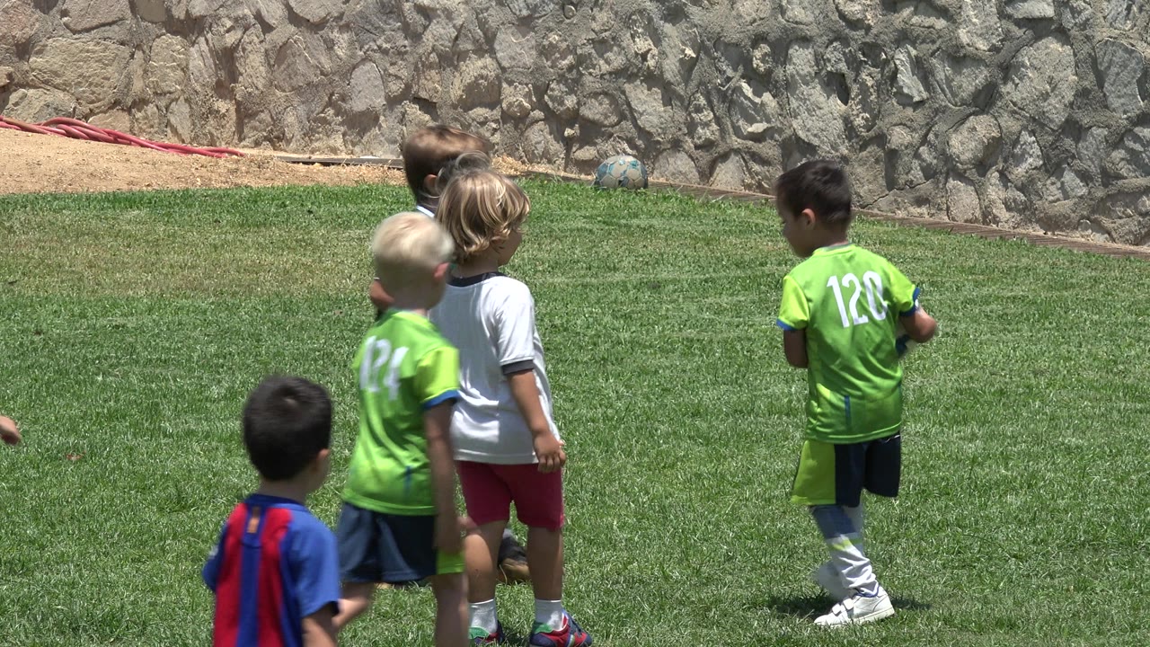 Young soccer players in Cabo San Lucas, Mexico stand and watch on the grass, with a ball near their feet and the stone wall filling the background.