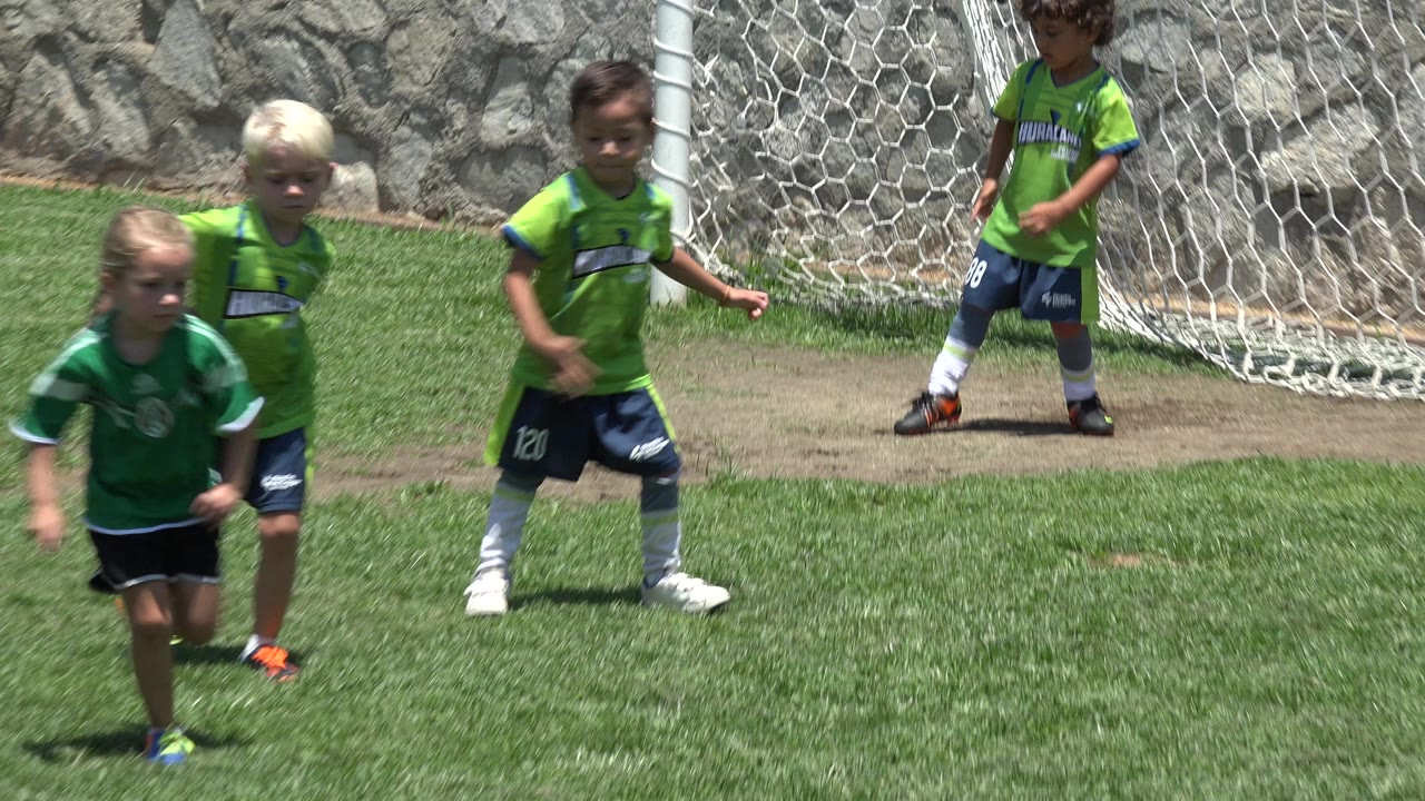 Several young players in green uniforms move across the grass in Cabo San Lucas, Mexico, while a white goal and chain-link fence sit behind them.