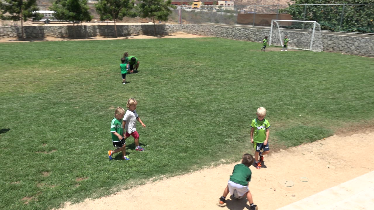 Young children in soccer shirts stand and crouch near the sideline of a grass field in Cabo San Lucas, Mexico, with a goal and stone wall in the background.