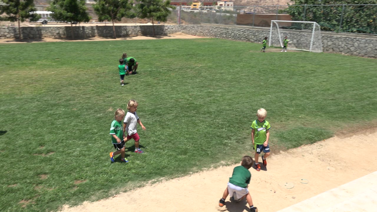 Children in green and white soccer clothing gather on the grass near a dirt edge in Cabo San Lucas, Mexico, while a small goal stands behind them.