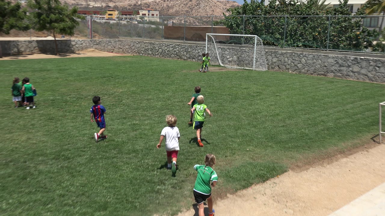 Young children in green, white, and blue shirts run across a grass soccer field in Cabo San Lucas, Mexico, with a white goal, stone wall, fence, and hillside buildings behind them.