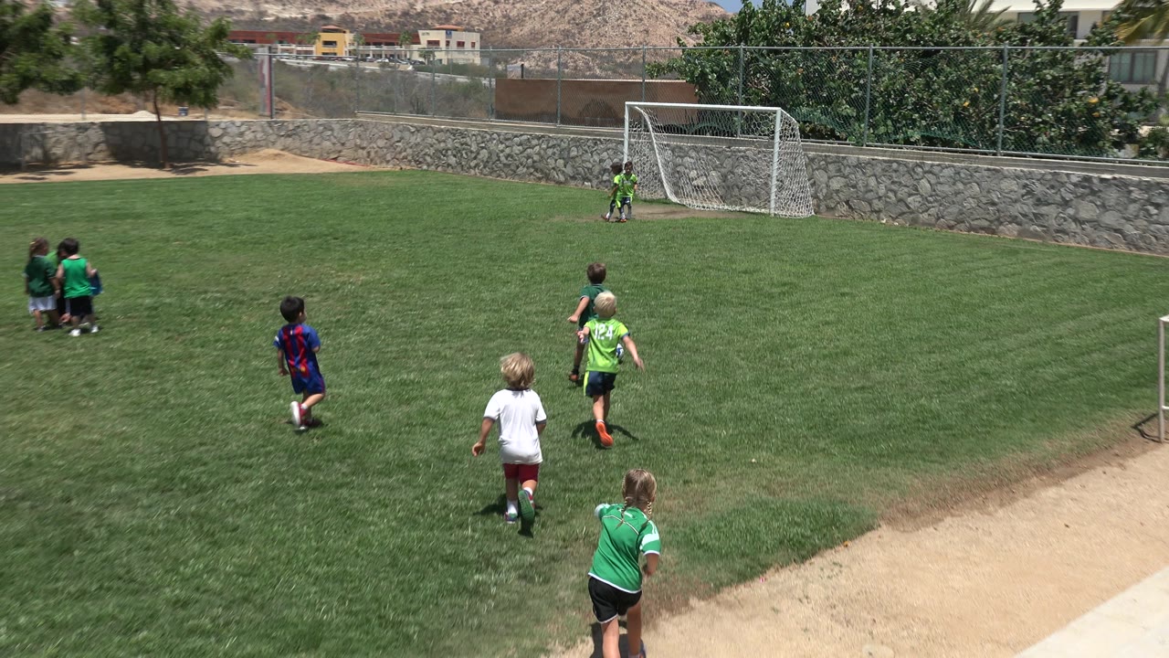 The children spread out across the grass field in Cabo San Lucas, Mexico, with one child in a blue and red shirt near the ball and a white soccer goal in the distance.