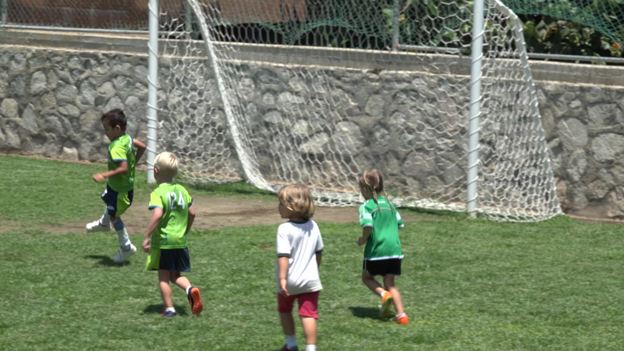 Several children move toward a light-colored soccer ball beside the goal net in Cabo San Lucas, Mexico, while another child in a white shirt watches from the field.