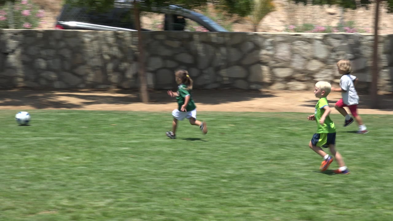 In Cabo San Lucas, the girl in green and white runs toward the soccer ball while two boys move behind her on the sunlit grass.
