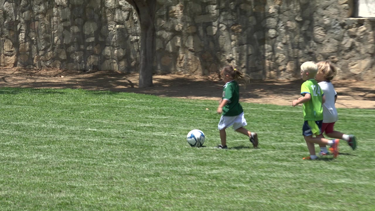 Three children move across the sunny grass toward the soccer ball in Cabo San Lucas, Mexico, with the stone wall and shaded dirt in the background.
