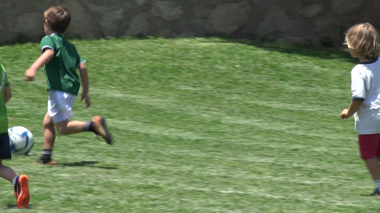 Children in green and white soccer uniforms run across a sunlit grass field in Cabo San Lucas, Mexico, with a soccer ball near the left edge and a stone wall behind them.