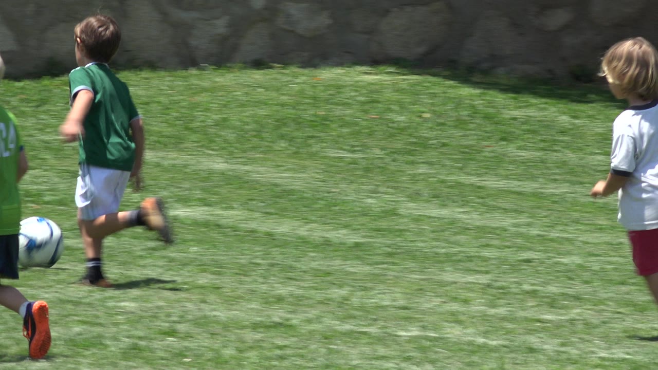Two children in soccer uniforms move near a ball on the grass in Cabo San Lucas, Mexico, with bright sun on the field and a stone wall in the background.