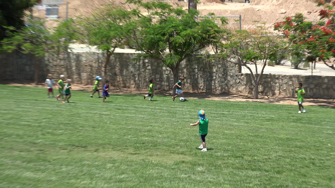 A child in a green shirt and blue cap stands in the foreground as other children move with a soccer ball near the stone wall in Cabo San Lucas.