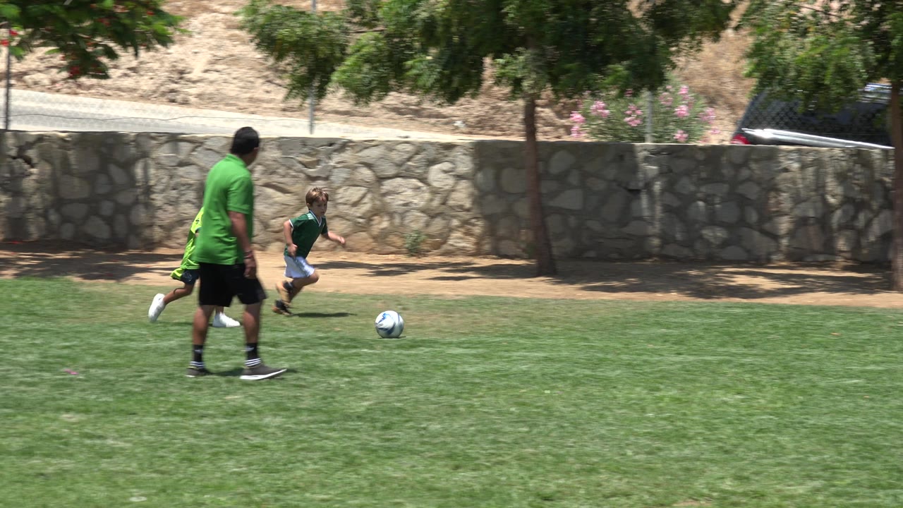 Two children in green soccer jerseys run toward a blue and white soccer ball while an adult trails behind on the grass field in Cabo San Lucas, Mexico.