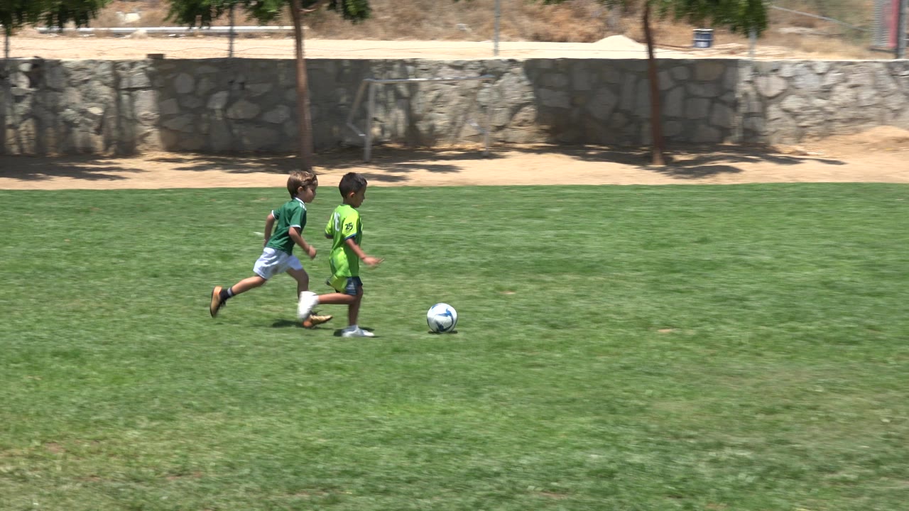 At the start of the Cabo San Lucas clip, two children in green jerseys chase a blue and white soccer ball across a sunny grass field.