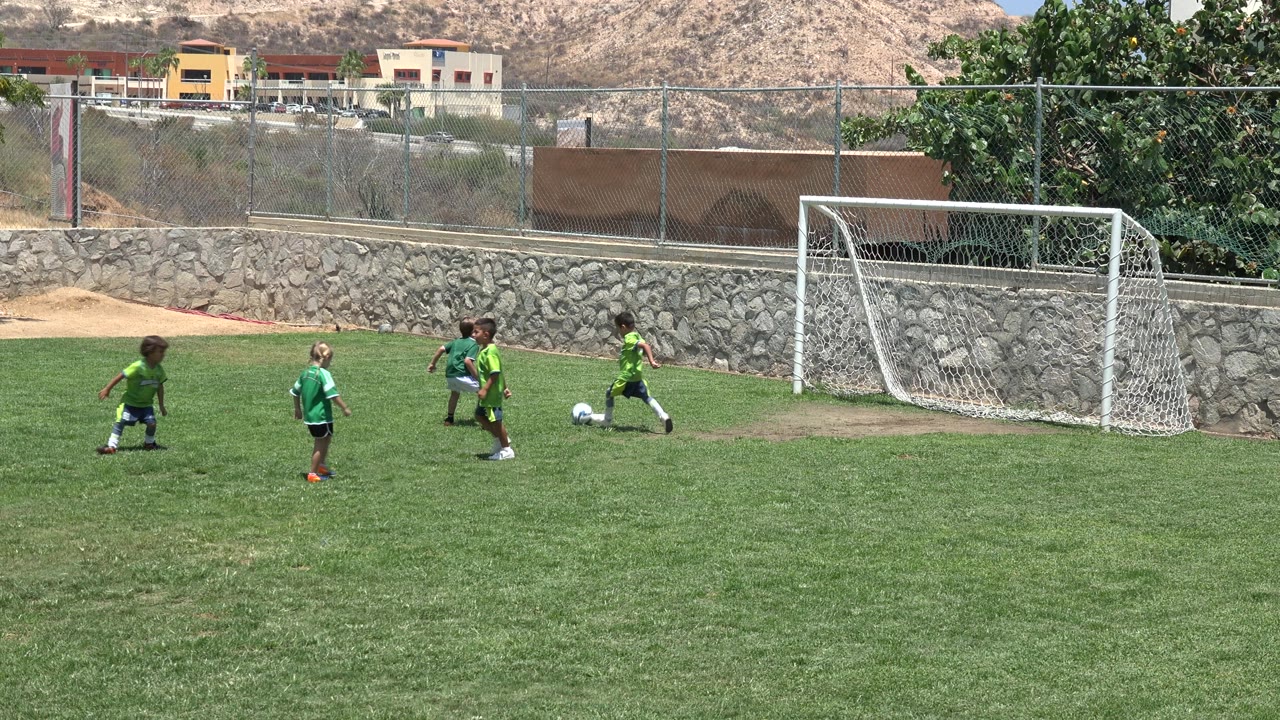 Children in green soccer jerseys play near a white goal on a sunny grass field in Cabo San Lucas, Mexico, with a stone wall and dry hillside behind them.