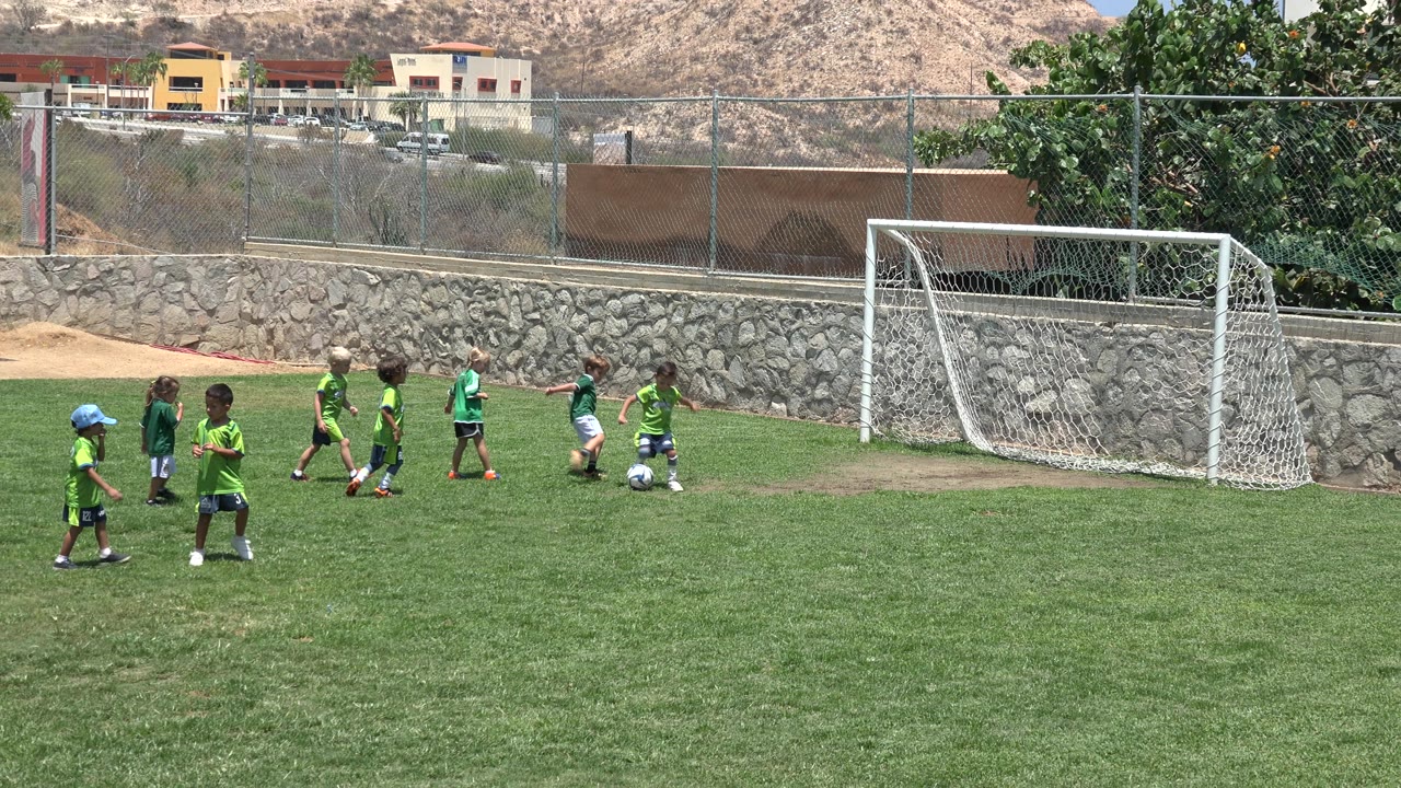 Children in green soccer uniforms gather and move near a white soccer goal on a grass field in Cabo San Lucas, Mexico, with a stone wall and hillside behind them.
