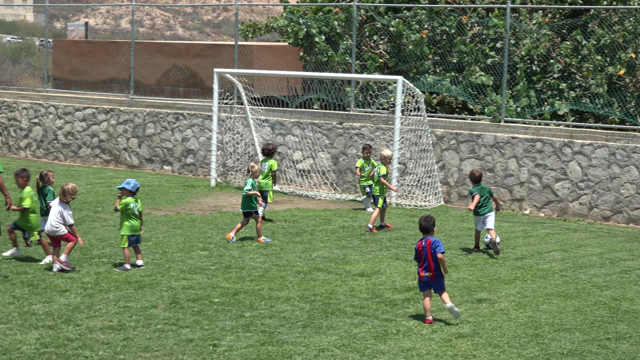 Several children in soccer clothing run toward a white goal on a sunny grass field in Cabo San Lucas, Mexico, with a stone wall and fence behind them.