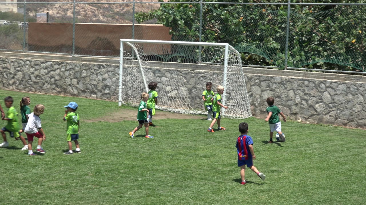 Children in green jerseys and shorts move across the grass toward the goal in Cabo San Lucas, Mexico, while another child in a blue and red shirt stands farther back.