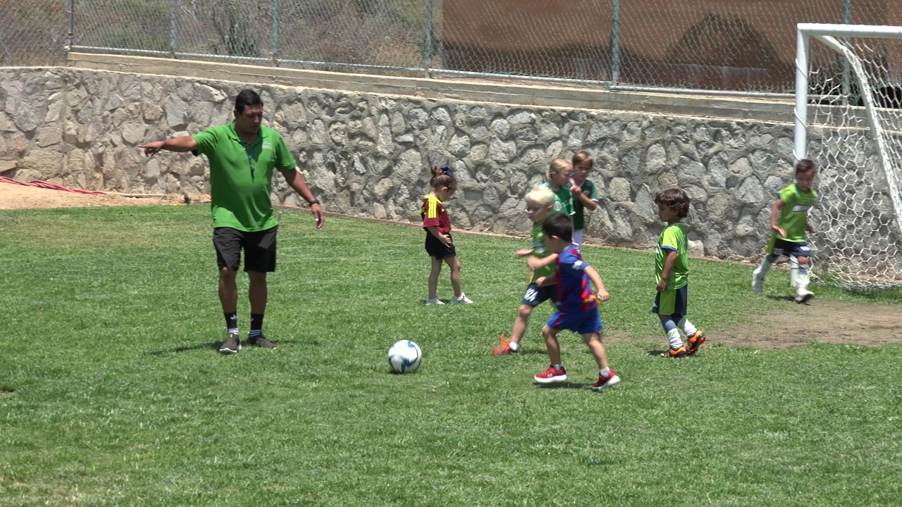 A coach in a green shirt points on a Cabo San Lucas soccer field while young children in uniforms gather near a ball and a white goal.
