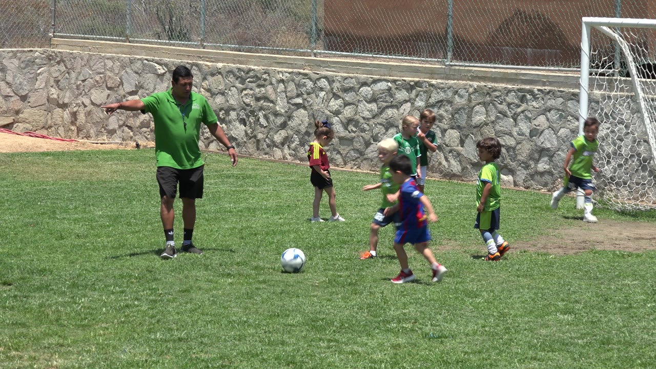 The coach remains near midfield in Cabo San Lucas as children in green and maroon soccer uniforms move around a ball in front of the goal.