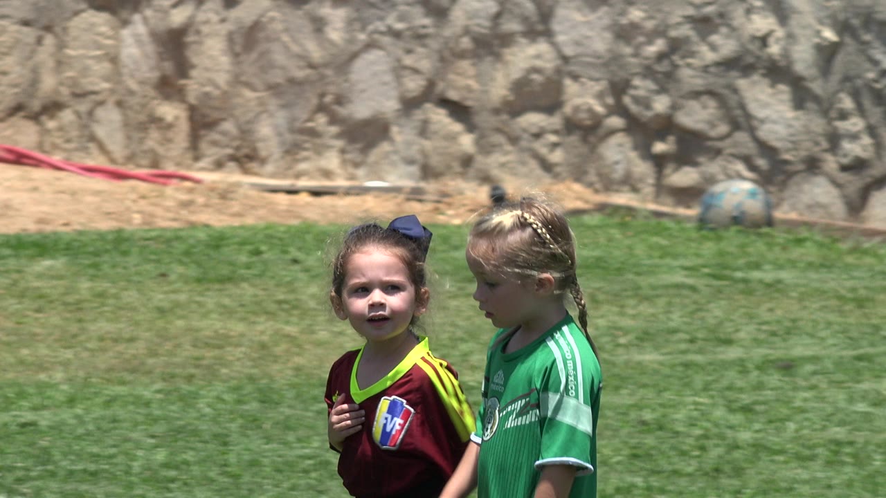 Two young girls in maroon and green soccer jerseys stand on grass in Cabo San Lucas, Mexico, with a rocky dirt bank behind them.