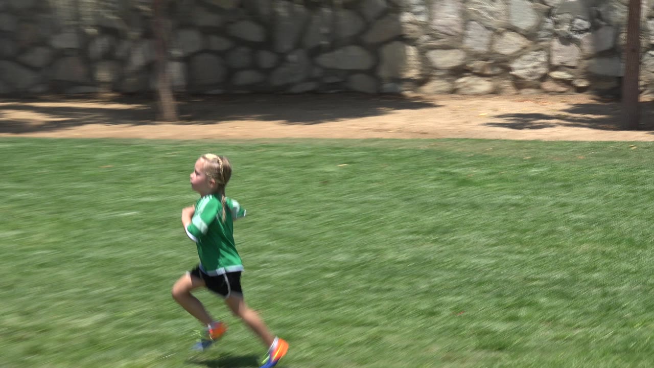 Child in a green soccer jersey running across a grass field.