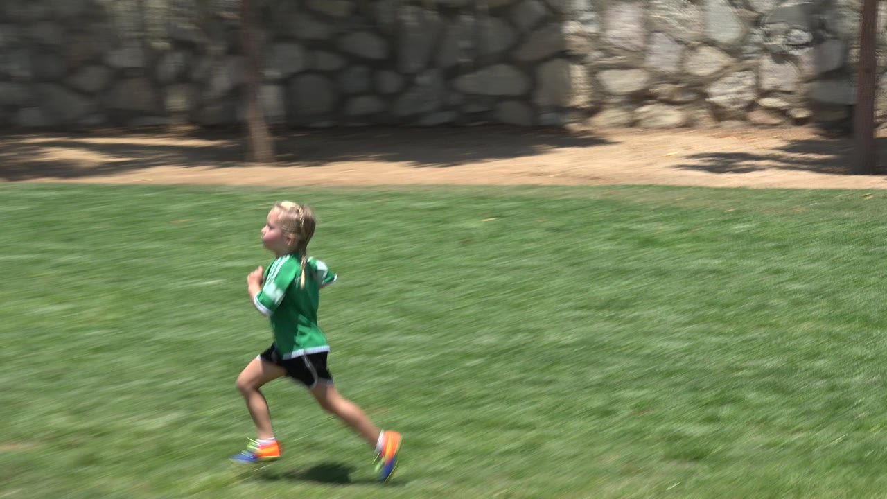 A young player in a green jersey crosses the grass field in Cabo San Lucas, Mexico, with the stone boundary wall and sunlit field filling the frame.
