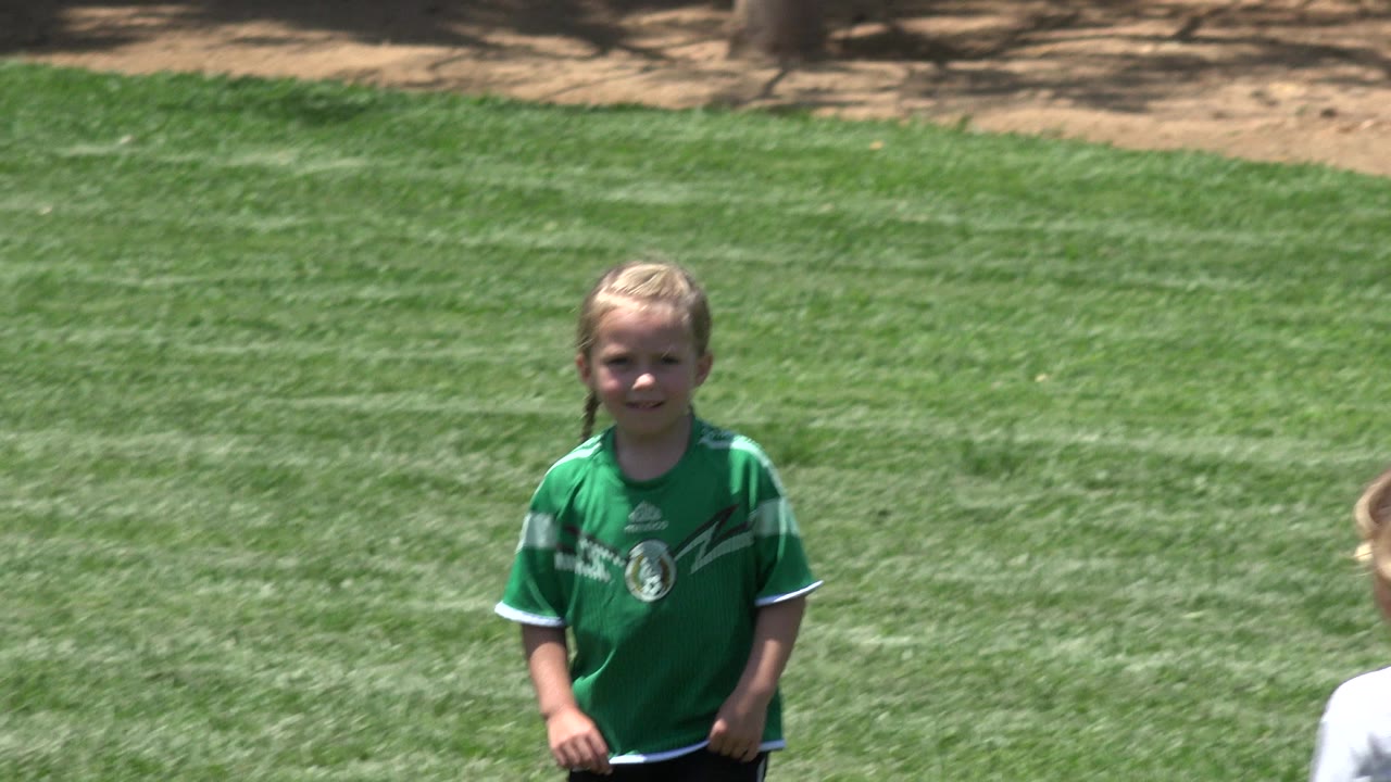 A young child in a green soccer jersey stands on a bright green lawn in Cabo San Lucas, Mexico, with a tan path area behind the field.