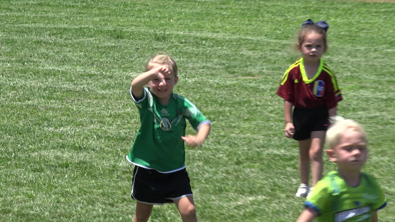 In Cabo San Lucas, a blonde child in a green shirt and black shorts walks near the foreground while another child in a burgundy shirt stands farther back on the grass.