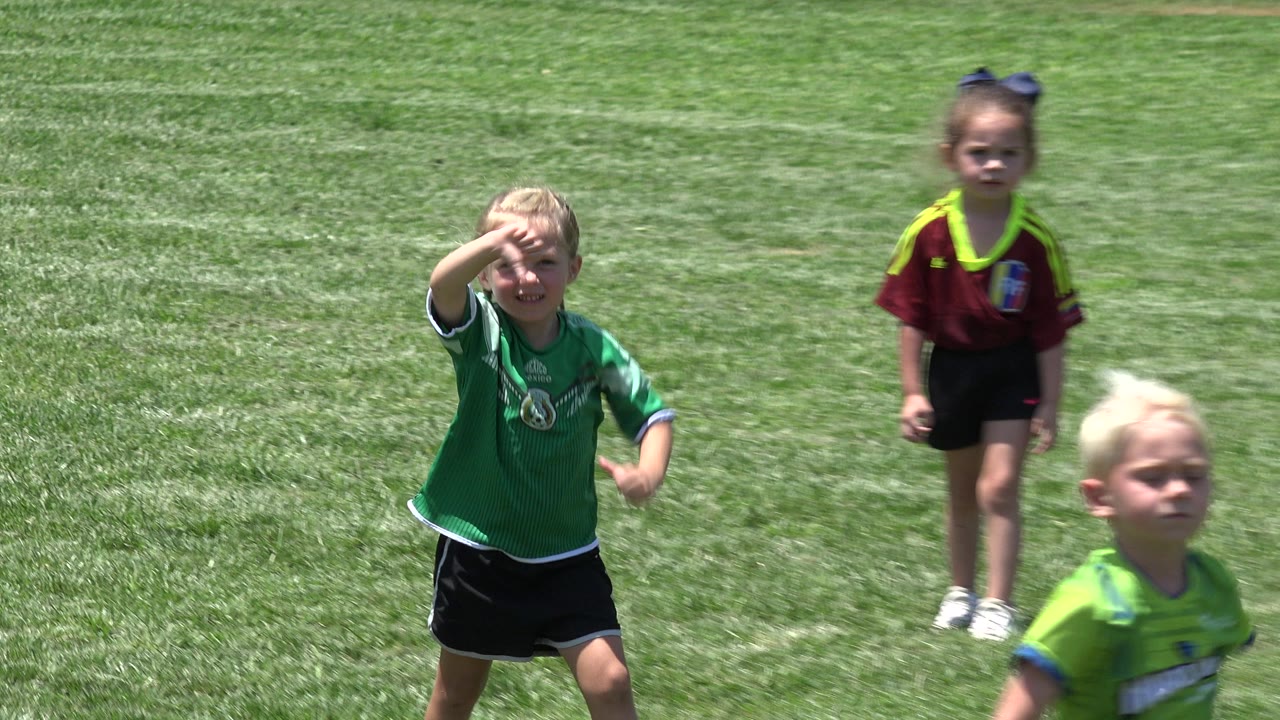 In Cabo San Lucas, the blonde girl in a green shirt raises a hand near her face as a child in a burgundy shirt and another child in green appear behind her on the lawn.