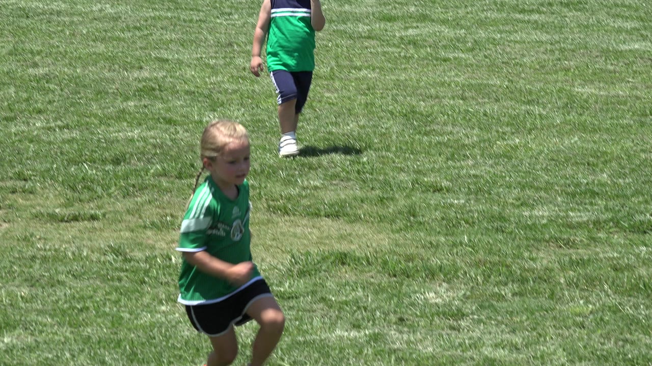 A young blonde girl in a green soccer jersey runs across a grassy field in Cabo San Lucas, Mexico, with another child partly visible behind her.