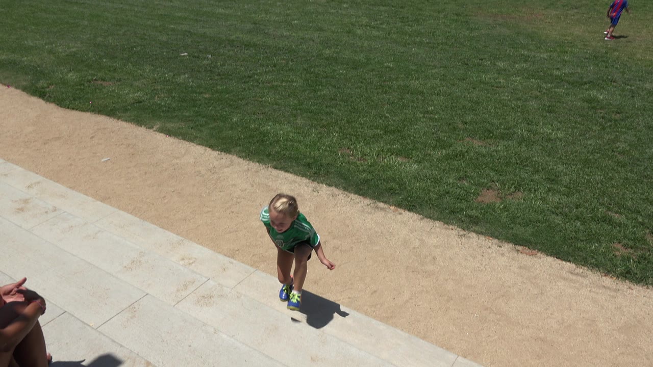 Child in green soccer jersey on a paved walkway beside grass and dirt.