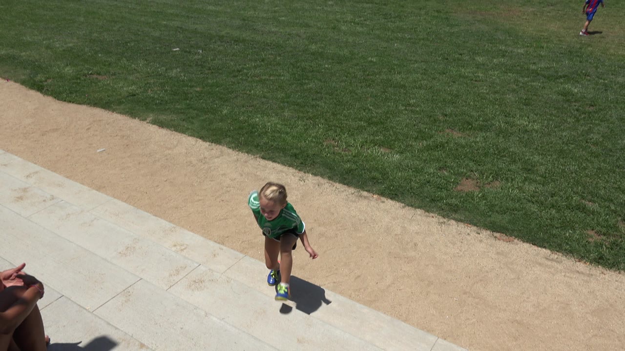 The child in the green jersey is centered farther along the paved edge, with the diagonal dirt path and green soccer field filling most of the Cabo San Lucas frame.