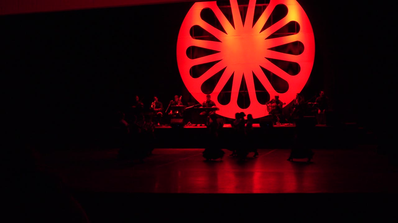 Dark stage with a large glowing red circular backdrop and musicians seated behind dancers.