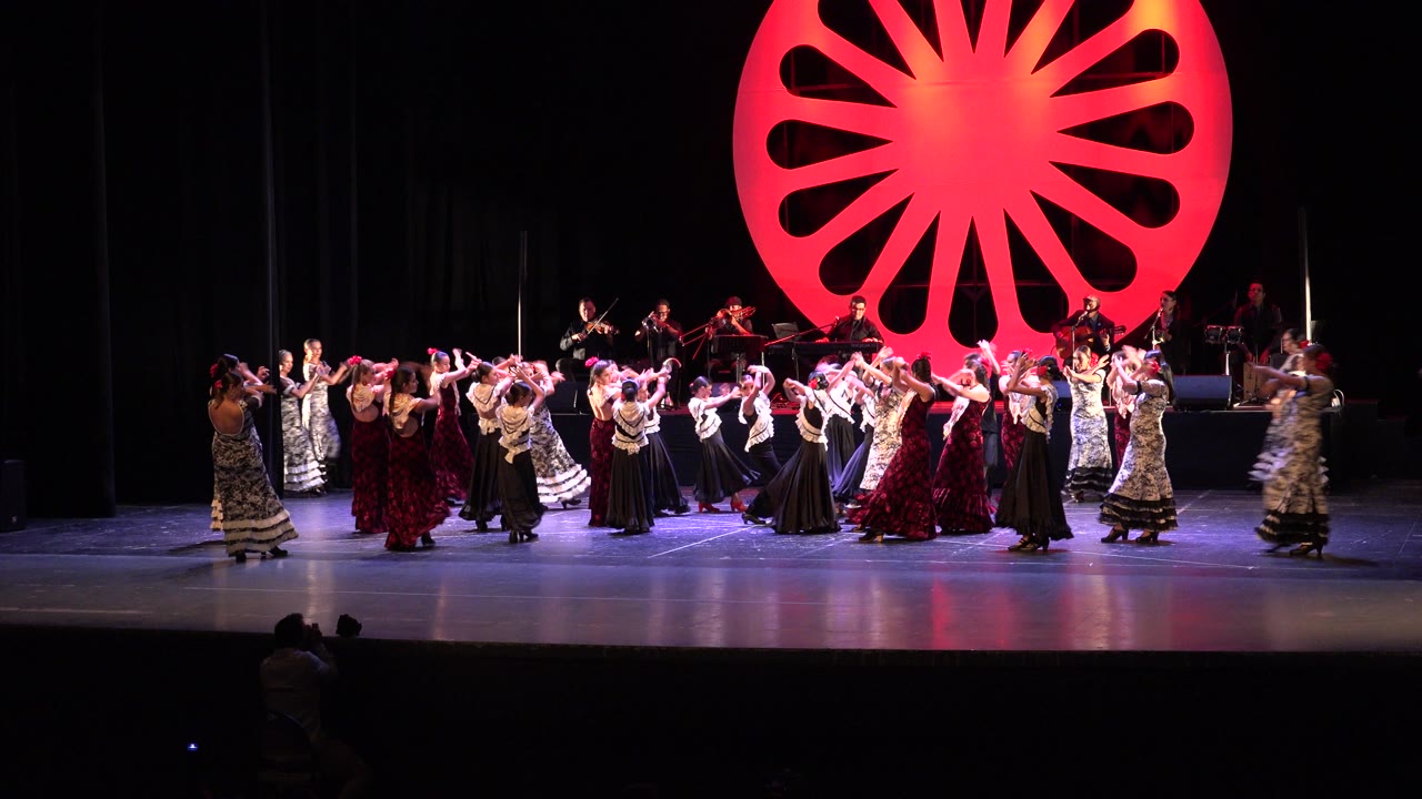 Wide view of a Cabo San Lucas stage show with many dancers spread across the stage, musicians behind them, and a large red circular set piece overhead.