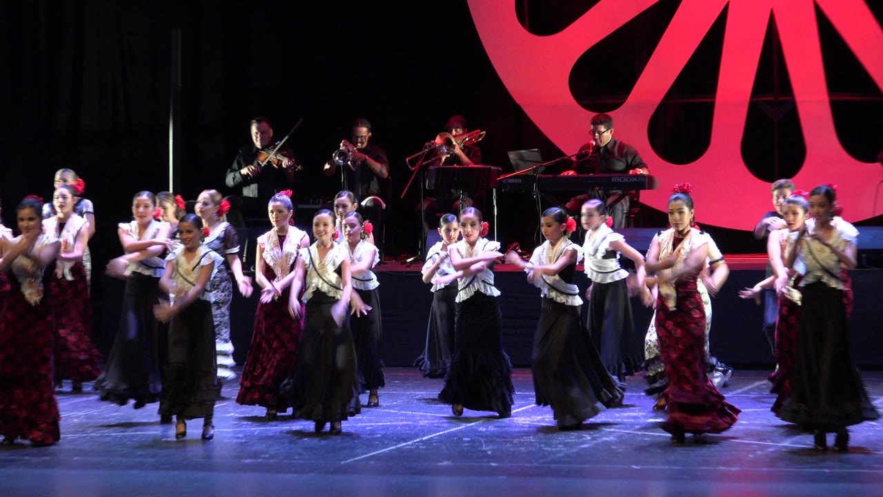 Young dancers continue a choreographed routine on a Cabo San Lucas stage, with arms crossed or extended and musicians visible across the rear platform.