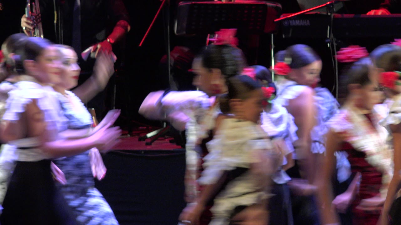 Girls in traditional-style costumes move across a dark stage in Cabo San Lucas, Mexico, with red hair flowers, white ruffles, and musical equipment behind them.