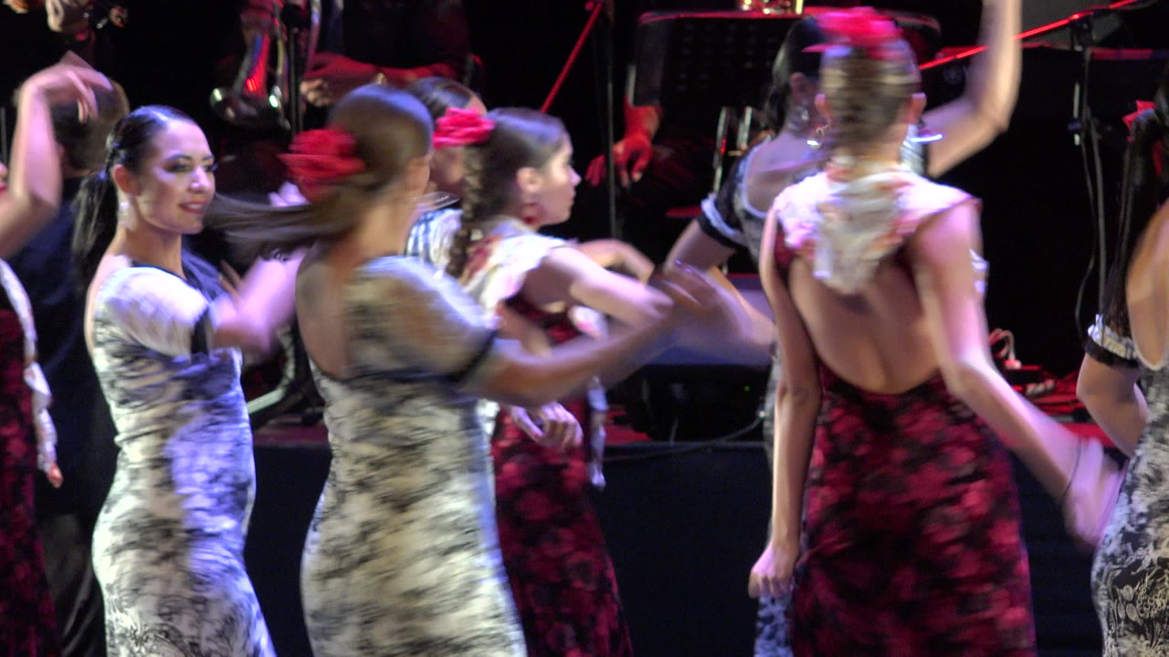 Women in red, white, and black patterned dresses dance on a stage in Cabo San Lucas, Mexico, with a live band partly visible in the dark background.